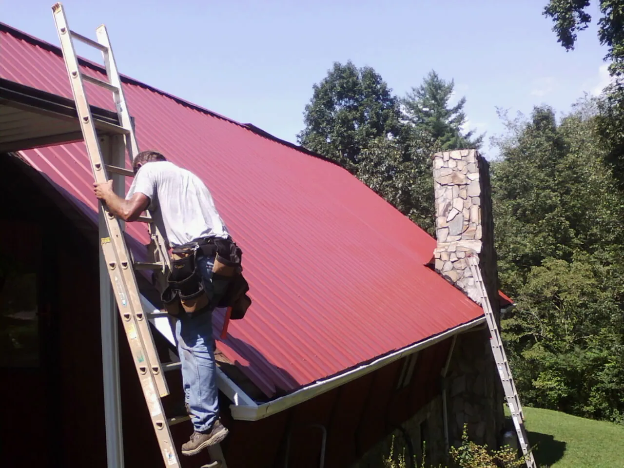 Roofer on a ladder beside a red metal roof and stone chimney — Soffit & Fascia Repair by Roofing Brothers in Stanhope